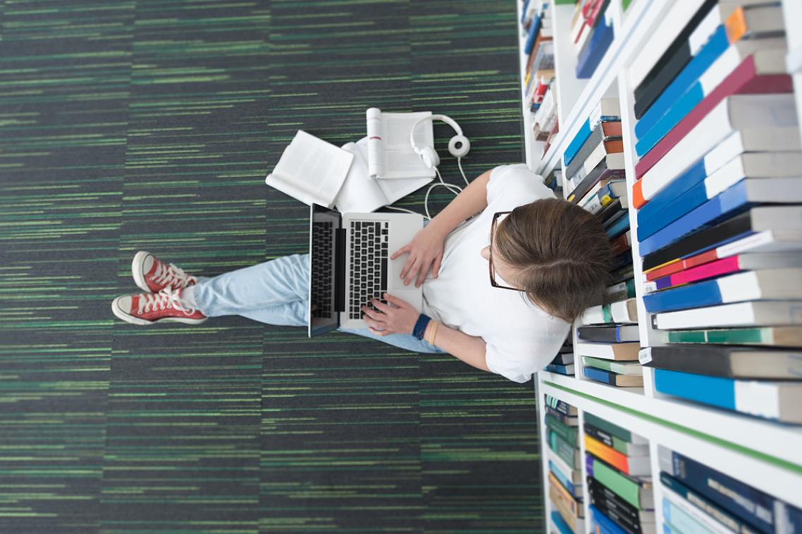 a student sits cross-legged on the floor of a library, typing on a laptop